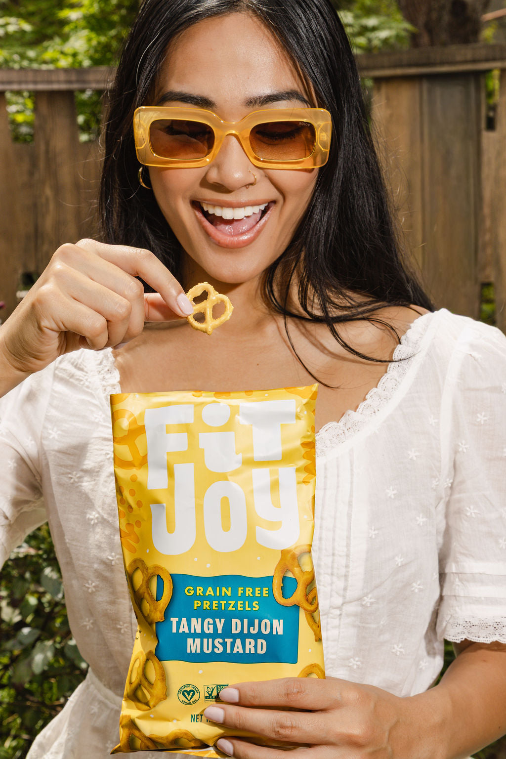 Smiling person outdoors holding a bag of FitJoy Tangy Dijon Mustard grain-free pretzels and lifting a pretzel twist